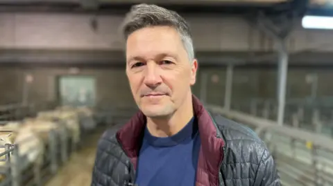 BBC John Whitby, wearing a black puffer jacket and blue t-shirt, looks at the camera. In the background, out of focus, there is a livestock market with some metal fences and sheep.