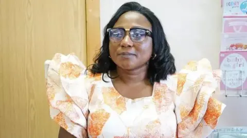 MEN Media Alberta Obinim, 43, has dark hair and wears glasses. She is wearing an orange and white dress – and is standing in front of birthday cards 