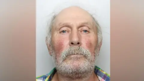 South Yorkshire Police A police mugshot of Schwalbe, who is facing directly toward the camera. He has very little hair on the top of the head, with some light-coloured hair remaining around the sides. A full white beard and moustache cover the lower face. He is wearing a multi-coloured plaid shirt with green, blue, yellow, and purple tones visible near the collar and shoulders.