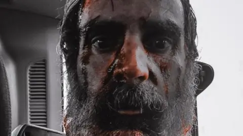 DJ Aerial Photography A close-up of a man with staring eyes and facial hair. His head is smeared in blood.
