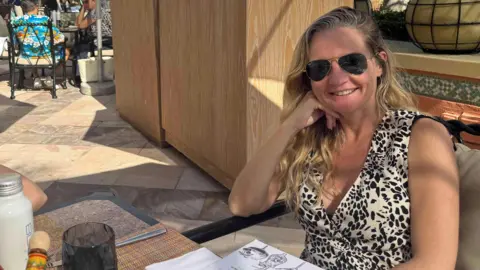 The image shows an outdoor restaurant or café area on a sunny day. In the foreground there is a person with long fair hair and wearing dark glasses seated at a table, along with outdoor furniture and decorative features.