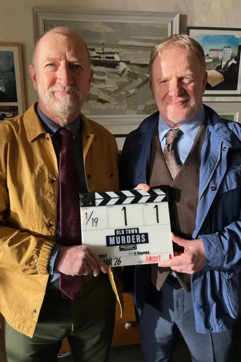Quay Street Productions Steffan Rhodri and Mark Lewis Jones holding a clapperboard between them. Rhodri is on the left in  a mustard jacket and a burgundy tie, with a blue shirt, while Jones is on the right wearing a blue jacket, blue shirt and brown waistcoat and tie. They are both stood up. 
