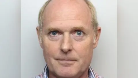 A custody picture of an older man with thinning hair who wears a checked shirt.