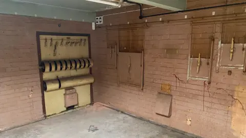 There is old projector hatches and a lighting rig pictured in the image. There is a grey concrete floor with light cream coloured tiled walls. 