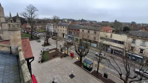 Arial view of Sleaford town centre. There is a row of three-storey buildings, with businesses occupying the lower floors. Cars are also visible on the road, with trees and a bus shelter in the foreground.