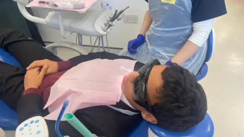 A patient is lays in a dental chair beside a dental nurse 