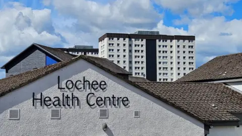 Lochee Health Centre which is a long, white, single-storey building with a slate rood. There are flats and houses around it and cars in a car park at the front and side.