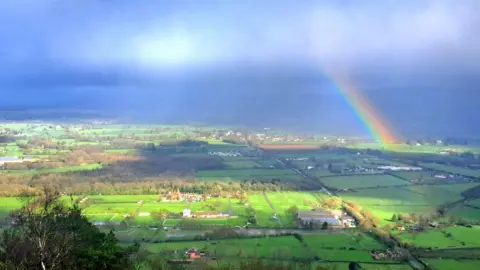 Blue skies with wispy cloud over the Malvern countryside. Some sunshine is peaking through but a vivid rainbow can also be seen on the left. 