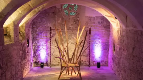 Changing Lives A large wooden chair which is made up of planks of wood of varying heights. It is placed in the centre of a stone room with a curved ceiling. There is a small, circular stained glass window at the top of the central wall. There are purple lights on either side of the char.