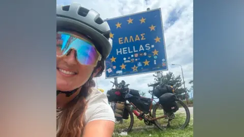 Matilda Pine Matilda Pine kneels and takes a photo of a sign behind her saying Hellas (Greece). The square sign is painted in European Union flag colours- blue with gold stars. Her bike with its carry-bags is propped up agaianst the sign.