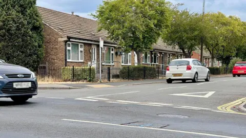 A crossroads with a row of bungalows. Three cars are on the road. In the middle of the crossroads is a manhole with a blue cross spray painted on it.