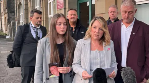 Natalie, wearing a grey pin strip business suit looks up as she speaks from a sheet of paper. A man is next to her in a suit and holding his head down. A young woman is to her right, holding a photograph of Callum in one hand