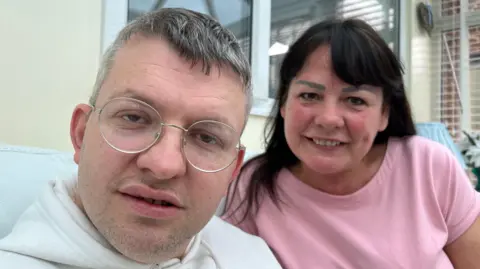 Leon Atkinson wears a cream hoodie and round gold rimmed glasses. He has salt and pepper coloured short hair. Behind him sits his mum Debra Dakeyne who has long dark, straight hair and is wearing a pink T-shirt.