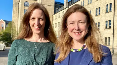 Green Party Head and shoulders shot of Catherine Braun wearing a green dress and Chloe Turner wearing a blue top and jacket looking at the camera standing in front of a former mill
