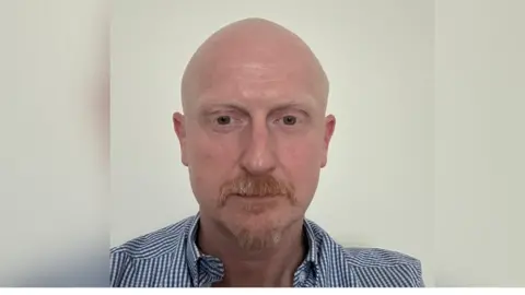 Brian McDaid Brian McDaid is bald with a ginger and grey moustache and goatee beard. He is wearing a stripey shirt and is looking down.