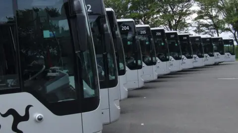 A row of buses lined up in a curved formation showing the driver's window in each. They are silver with dark tinted windscreens.