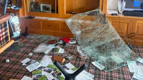 Significant damage to a wooden display cabinet following an apparent impact, with shattered glass panels, debris scattered across the floor, and contents displaced throughout the room.