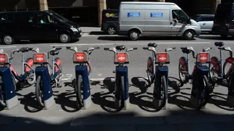 A row of seven Santander Cycles bicycles are parked in docking stations, viewed from the front on. The front of the bicycles display a red sign with the Santander Cycle and Transport for London roundel. The bikes are parked on the edge of the pavement, alongside a road, with vans and other traffic visible in the background.