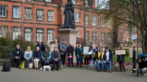 LDRS A group of around 18 people standing around a statue, which is outside a large Victorian redbrick building. Some are holding placards.