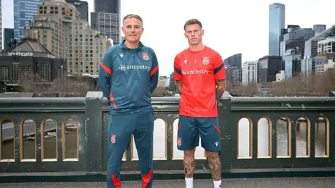 Phil Parkinson stood in a petrol-coloured Wrexham tracksuit with arms behind back next to James McClean in shorts and red training top. They are stood on a bridge with the Melbourne cityscape behind them. 