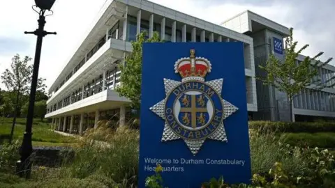 Durham Constabulary Headquarters, which is a modern rectangular building with a lot of windows. A large sign with force's crest on it is at the entrance.