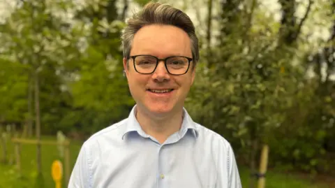 Cameron Watt outside with trees and grass behind him. He wears a light blue shirt and glasses. He has greying hair.