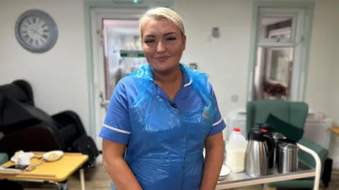 Sarah Clarke, who is wearing a blue healthcare tunic and an apron, is smiling at the camera in the dining room. There is a car with tea and coffee behind her. 