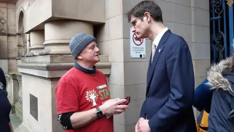 Julia Armstrong/BBC A man with a grey beanie hat and a red t-shirt talks to a taller bespectacled man in a navy suit. They are both stood outside a Town Hall.