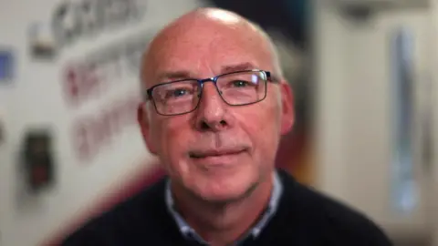 A balding man in his 60s with thin black glasses, wearing a light blue shirt and dark blue jumper