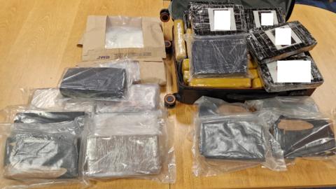 Parcels of cocaine, wrapped in black paper and placed in clear plastic bags, displayed on wooden table.