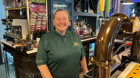 Helen Smith smiling at the camera. She is wearing a green t-shirt and is stood behind the bar in a pub.