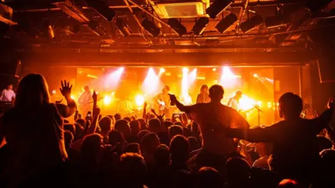 A general view from the back of the Leadmill music venue. A man, Miles kane, is on stage in the distance and a crowd with people dancing and sitting on each others shoulders is lit under orange lights