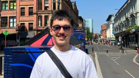 BBC/@harryadavis Harry in the foreground wearing a white T-shirt and sunglasses, with a black crossbody bag, standing on a pavement beside a blue-painted utility structure. Behind, a two-lane street has white road markings and a separated section with short black bollards. Pedestrians walk along pavements on both sides. Red-brick and light-coloured buildings line the street, with shopfronts and signage visible. A lamppost stands above the pavement, and taller modern buildings appear in the distance under a clear blue sky.