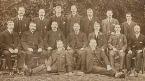 A black-and-white archival photo was provided showing 16 men wearing dark formal suits. Seven are standing in the back row, seven are sitting in the middle row, and two are lying on the ground in the front.