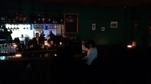 A bar in darkness because of the power cut, with people sitting at candlelit tables.