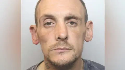 A mugshot of a bald man with a moustache and beard. He has a tattoo on his neck and is wearing a dark grey t-shirt. 