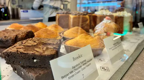 Cakes at Kacao. We can see a Brookie with belgian milk chocolate and white chocolate. There are also a range of pastries. In the background we can see someone working but it is blurred.