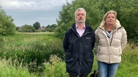 Two people stand side‑by‑side in outdoor jackets beside a grassy, pond‑like area with tall reeds, trees, and distant houses under a cloudy sky.