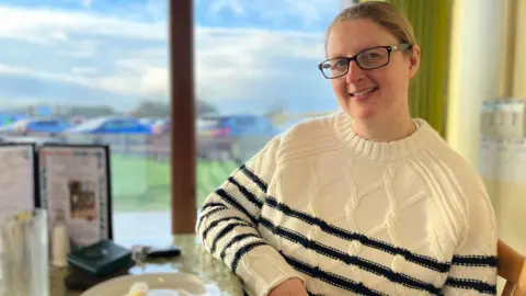 Shiona Bickley sitting at a table at the cafe at Watchtree Nature Reserve near Carlisle. She has blonde hair tied in a ponytail and wears glasses with a dark rim and a white knitted jumper with dark stripes.