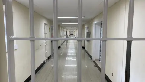 The camera looks down a long prison corridor, with a grey lino floor and what appear to be doors, with bars across them. There is a grey barred door in the front of the image, blocking the photographer's access.