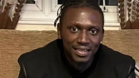 Hertfordshire Police Mustapha Jatta, smiling a the camera and wearing a black jacket. He is sitting on a sofa in a living room.