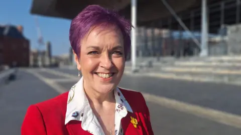Dawn Bowden outside the Senedd. She is in focus and the background is blurred. She smiles and has purple hair. She wears a red jacket and under a white patterned shirt. She has a daffodil on the left lapel. It is a head and shoulders shot.