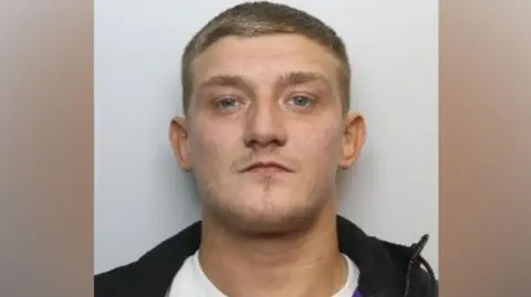 South Yorkshire Police A mugshot of Ivermee against a plain light grey background. He has very short, closely cropped hair and is wearing a white crew-neck T-shirt beneath a dark jacket. A section of purple fabric is visible inside the jacket near the right shoulder. The jacket is unzipped and sits open around the neck.