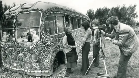 A bearded man in an anorak is looking through the viewfinder of a video camera at an elderly woman who is being interviewed on tape by a younger woman. A young man is watching on. They are standing beside a dilapidated bus which is covered in sea shells and has dolls in the windows.