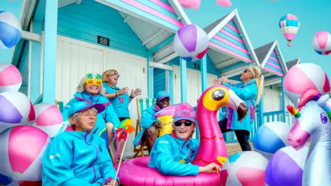 People wearing light blue jackets with bright coloured inflatable balls and a inflatable pink flamingo. There are colourful wooden huts behind them and a hot air balloon in the sky.