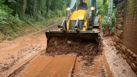 Travel Somerset A yellow digger removing thick mud from the road beneath a railway bridge.