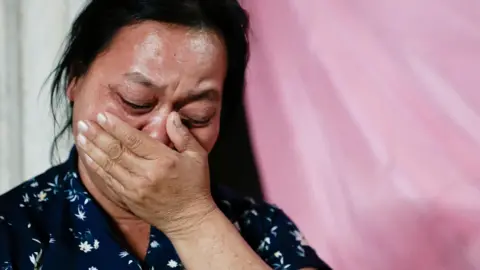 Reuters Wiwwaew Sriaoun, 53, mother of Thai hostage Watchara Sriaoun who was captured during the deadly October 7, 2023 attack by Hamas, holds her hand to her mouth, her eyes closed as as she weeps. She is wearing a dark blue blouse and stands against a pink background