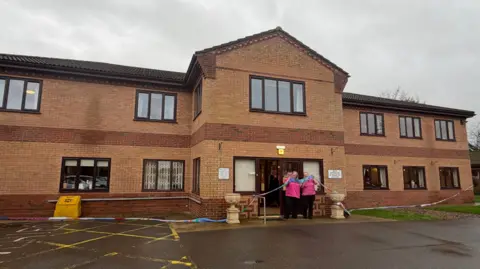 BBC/Amber Gash The finished scarf wrapped around the Tanglewood Care Home building. There are two members of staff dressed in pink holding it aloft at the entrance to the two-storey building.