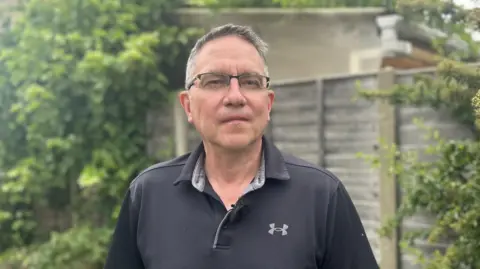 Kate Bradbrook/BBC A man with short grey hair is wearing glasses and a black polo shirt while standing in front of a garden fence.
