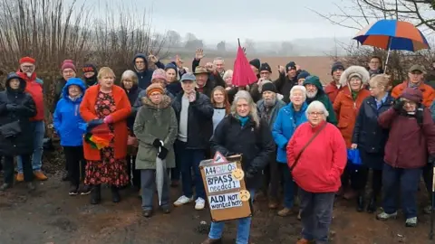 A large group of residents wearing coats and holding umbrellas are gathered by a brown field on a rainy day. One woman at the front of the group is holding a sign that reads: "A33 Chaos. Bypass needed. Aldi not wanted here." 